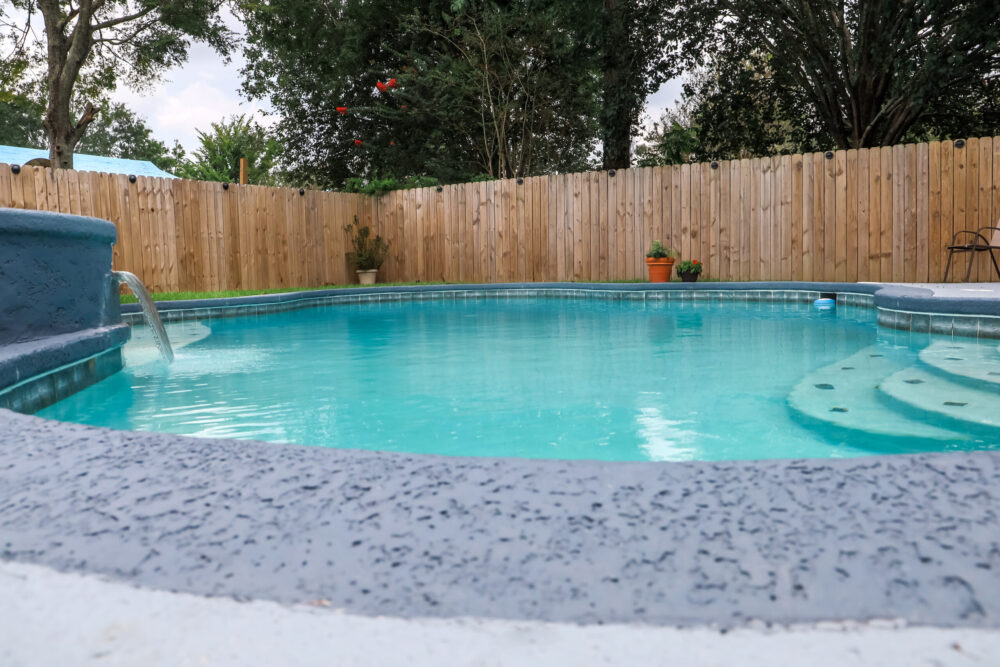 Low angle view of a large free-form gray grey accent swimming pool with turquoise blue water in a fenced-in backyard in a suburban neighborhood